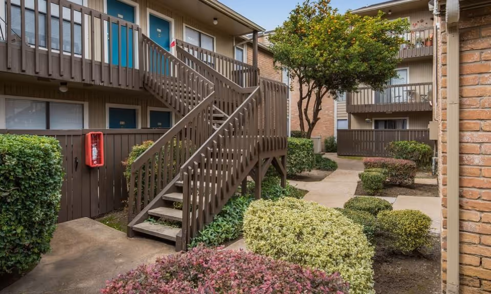Courtyard with wooden exterior staircases, balconies, doors and landscaped shrubs between apartment buildings.