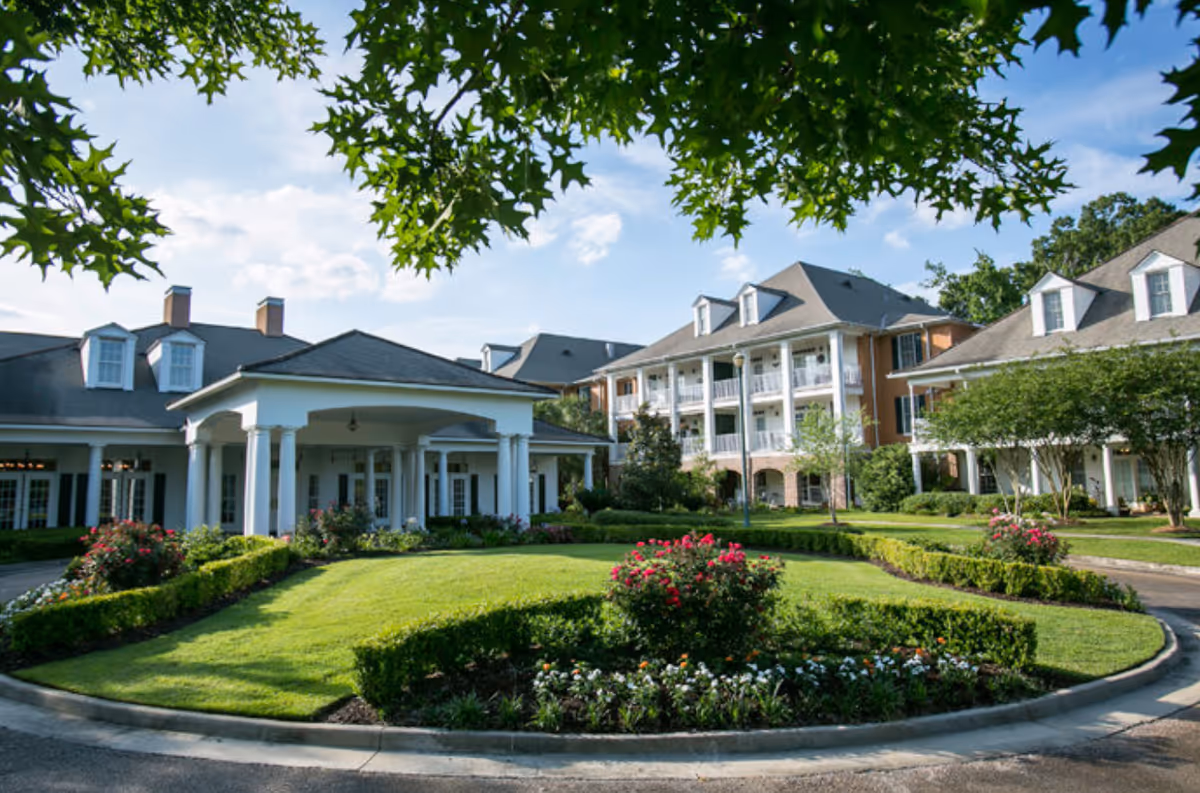 Exterior view of St. James Place senior living facility showing a well-maintained garden with green grass, flower beds, and bushes in the foreground. The building features white columns, multiple windows, balconies, and a gray roof under a partly cloudy sky. Tree branches frame the top of the image.