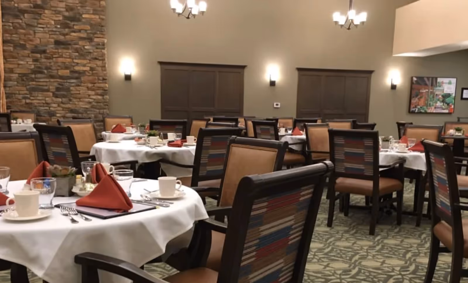 Dining room with round tables set for a meal, upholstered chairs, and warm wall and ceiling lighting.