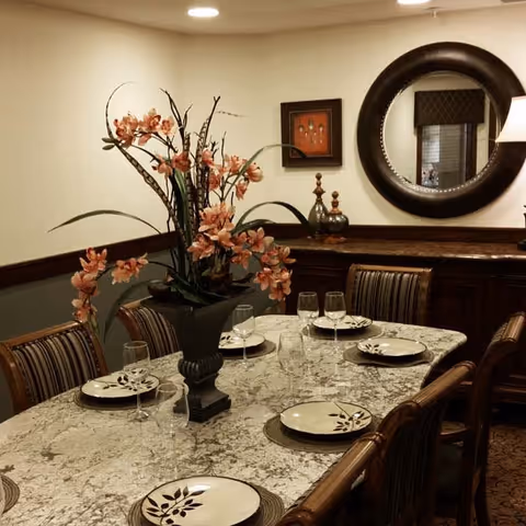 A dining room with a rectangular marble table set for six people with plates and wine glasses. A large floral arrangement in a black vase is centered on the table. The room features striped upholstered chairs, a wooden sideboard with decorative items, a round mirror on the wall, and a framed artwork.