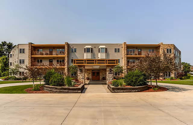 Front exterior view of a three-story senior living facility building with balconies, stone and beige siding, landscaped bushes and trees, and a paved driveway leading to the entrance.