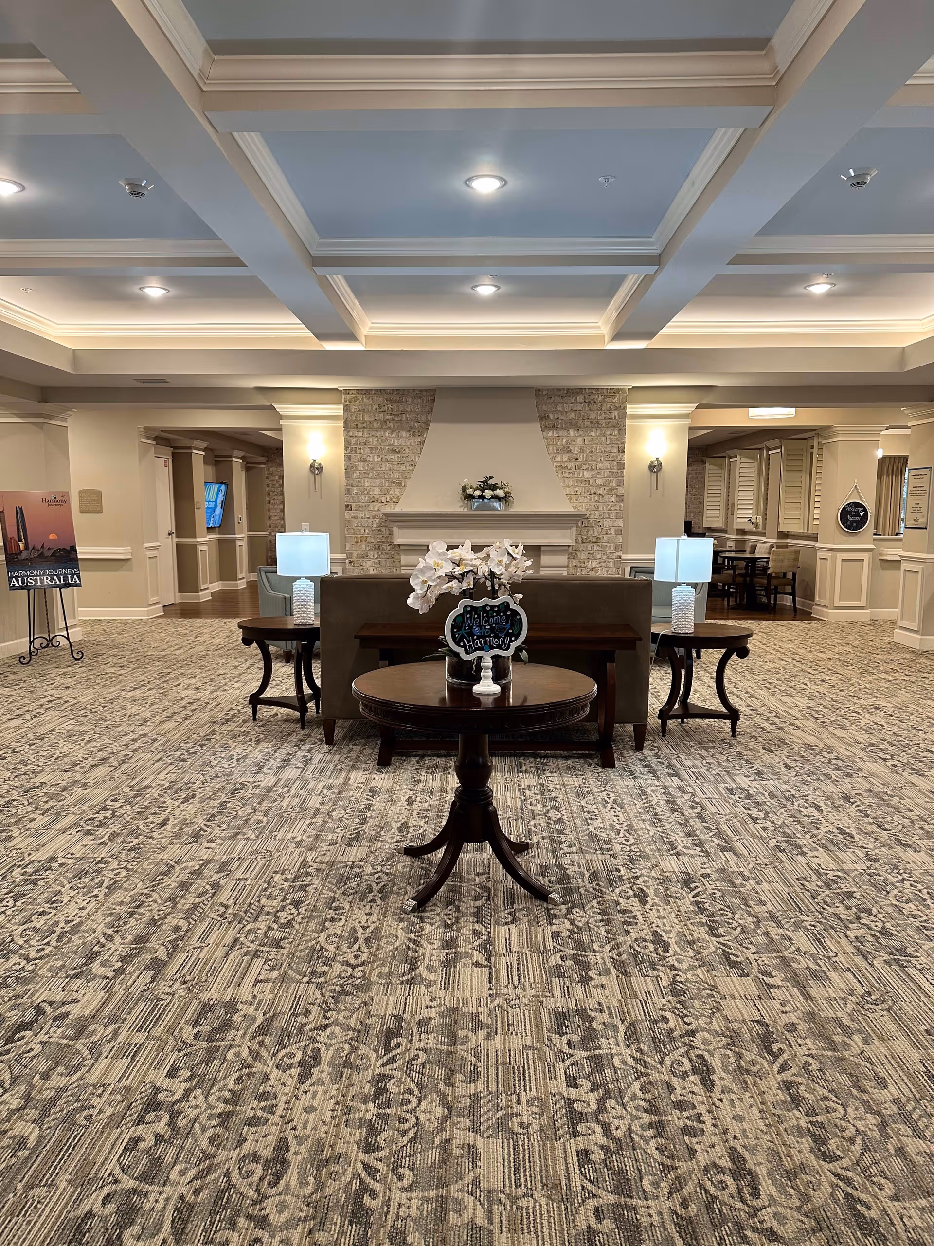Spacious senior living facility common area with patterned carpet, a central round wooden table with white orchids and a small sign that says 'Welcome to Harmony'. Behind the table is a sofa with two side tables each holding a lamp. The back wall features a brick fireplace with a flower arrangement on the mantel. The ceiling has recessed lighting and decorative beams. To the left and right are hallways and additional seating areas.
