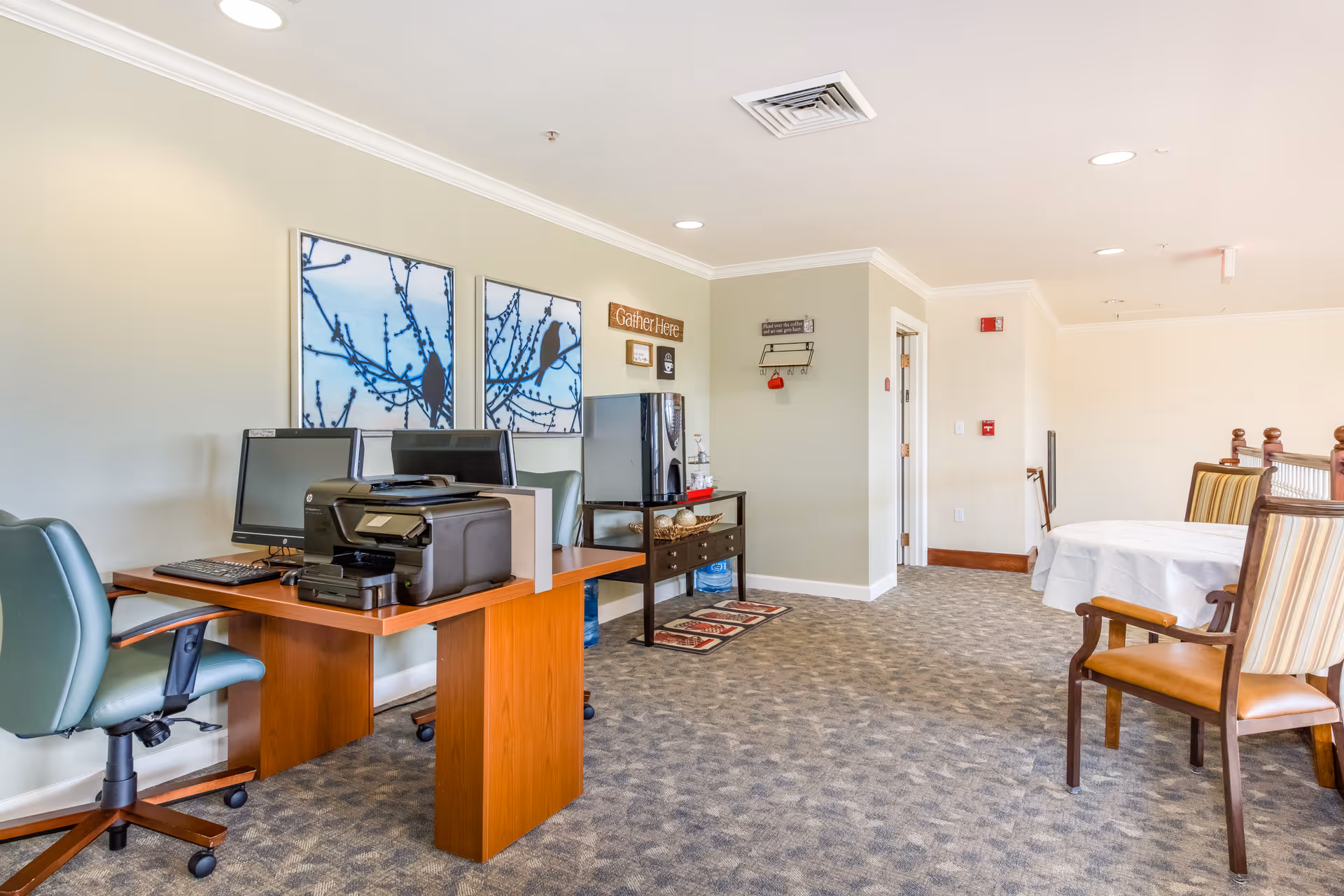 A bright communal interior with a desk workstation and printer on the left and a dining table with chairs on the right.