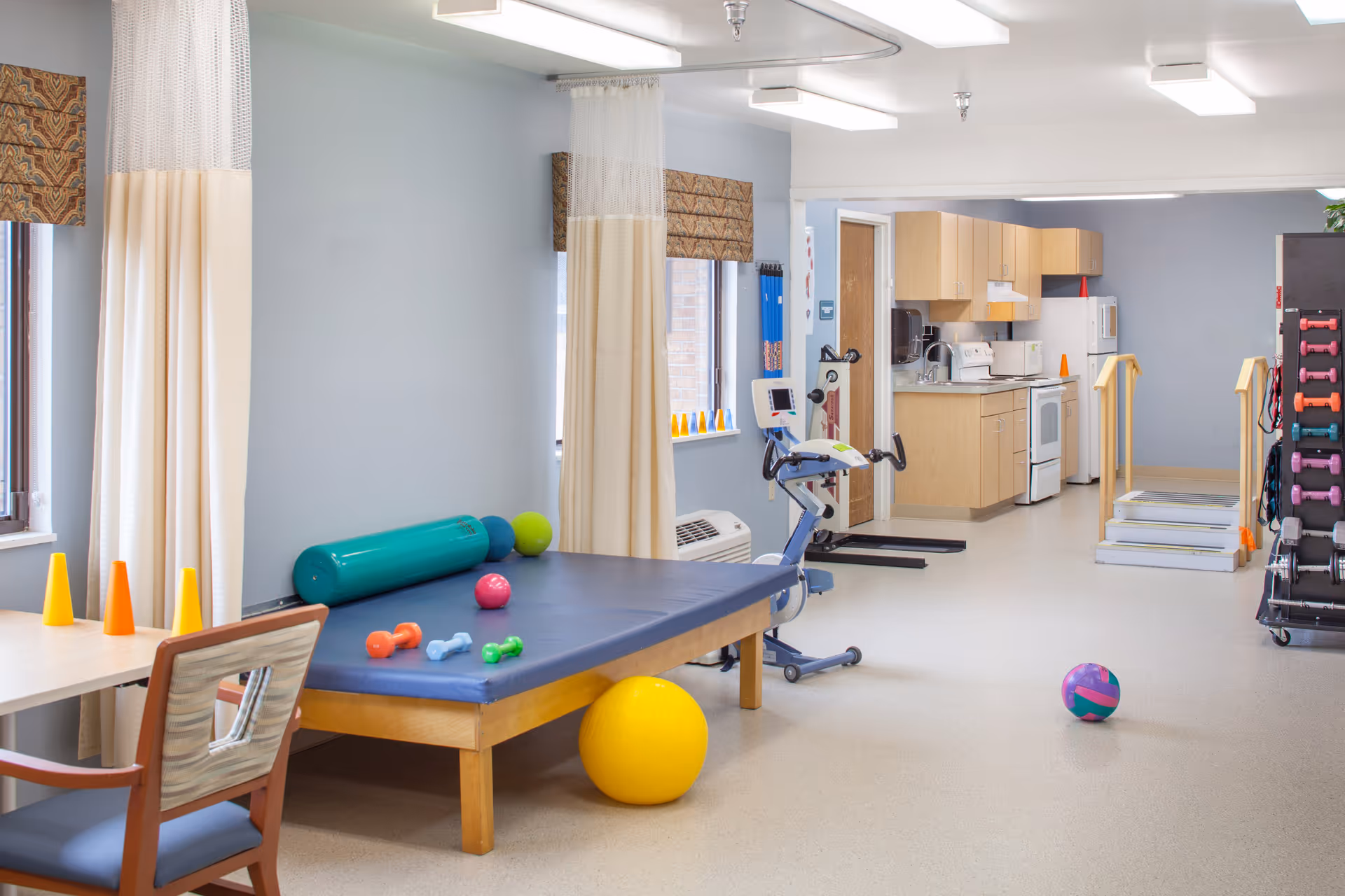 A bright therapy room with exercise equipment including a padded table with small dumbbells and exercise balls, an exercise bike, and a rack of colorful dumbbells. The room has light blue walls, windows with patterned valances and cream curtains, and a kitchenette area with wooden cabinets, a stove, microwave, and refrigerator. There are also handrails and steps for physical therapy exercises.