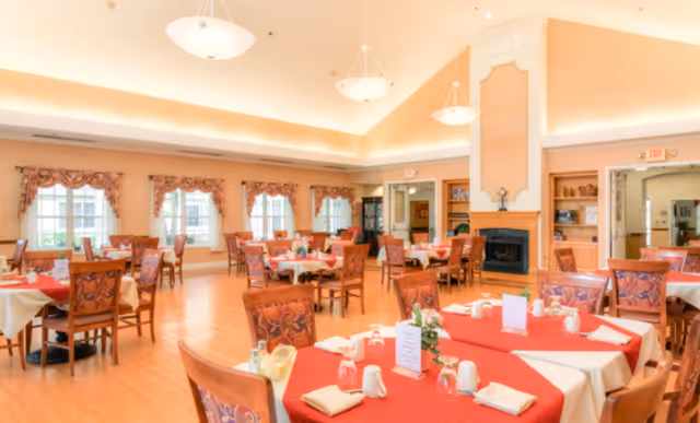 A spacious dining room with multiple round tables covered in red and white tablecloths, set with glasses, napkins, and menus. The room features wooden chairs with patterned upholstery, large windows with decorative curtains, a fireplace, and warm lighting from ceiling fixtures.