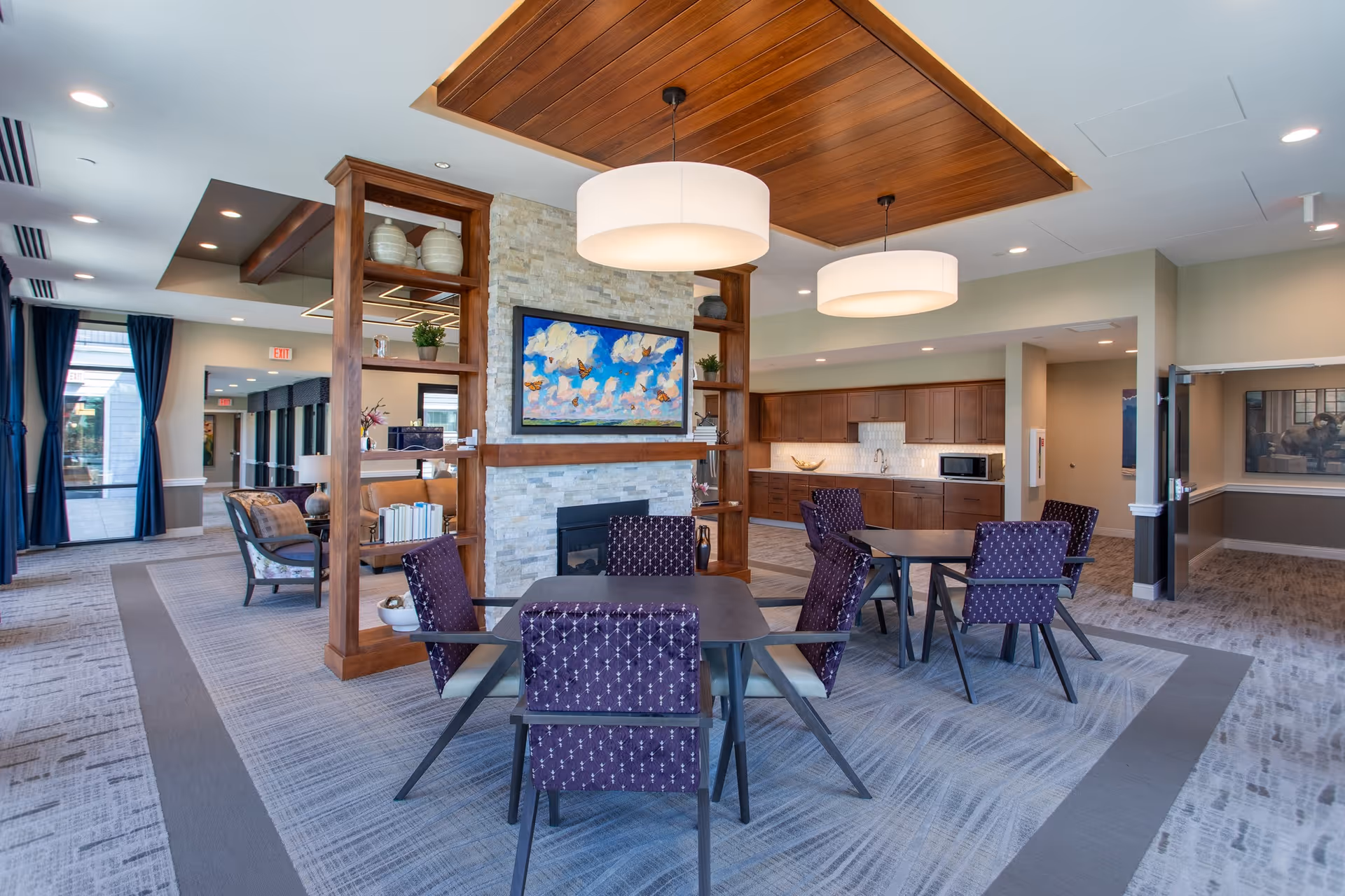 Communal senior living lounge with dining tables and purple chairs, a central stone fireplace with shelving, kitchenette, and pendant lights.