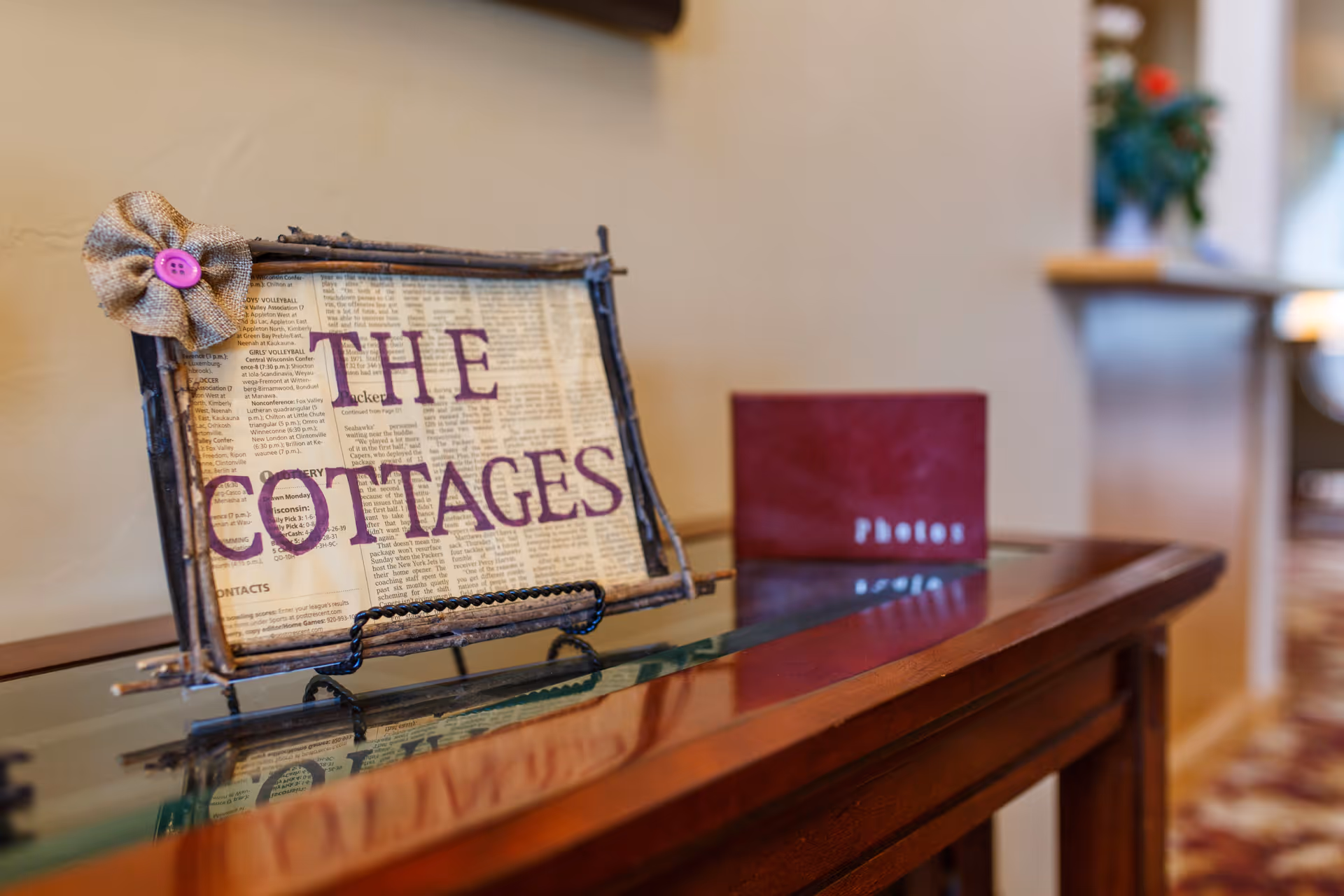 A decorative sign on a wooden table with a glass top that reads 'THE COTTAGES' in purple letters on a background resembling newspaper print, next to a red photo album labeled 'Photos'. The background shows a blurred interior space with a plant and furniture.