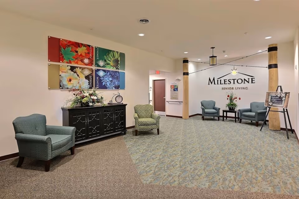 Lobby seating area of Milestone Senior Living with armchairs, a console topped with flowers, colorful wall art, and the facility logo on the far wall.