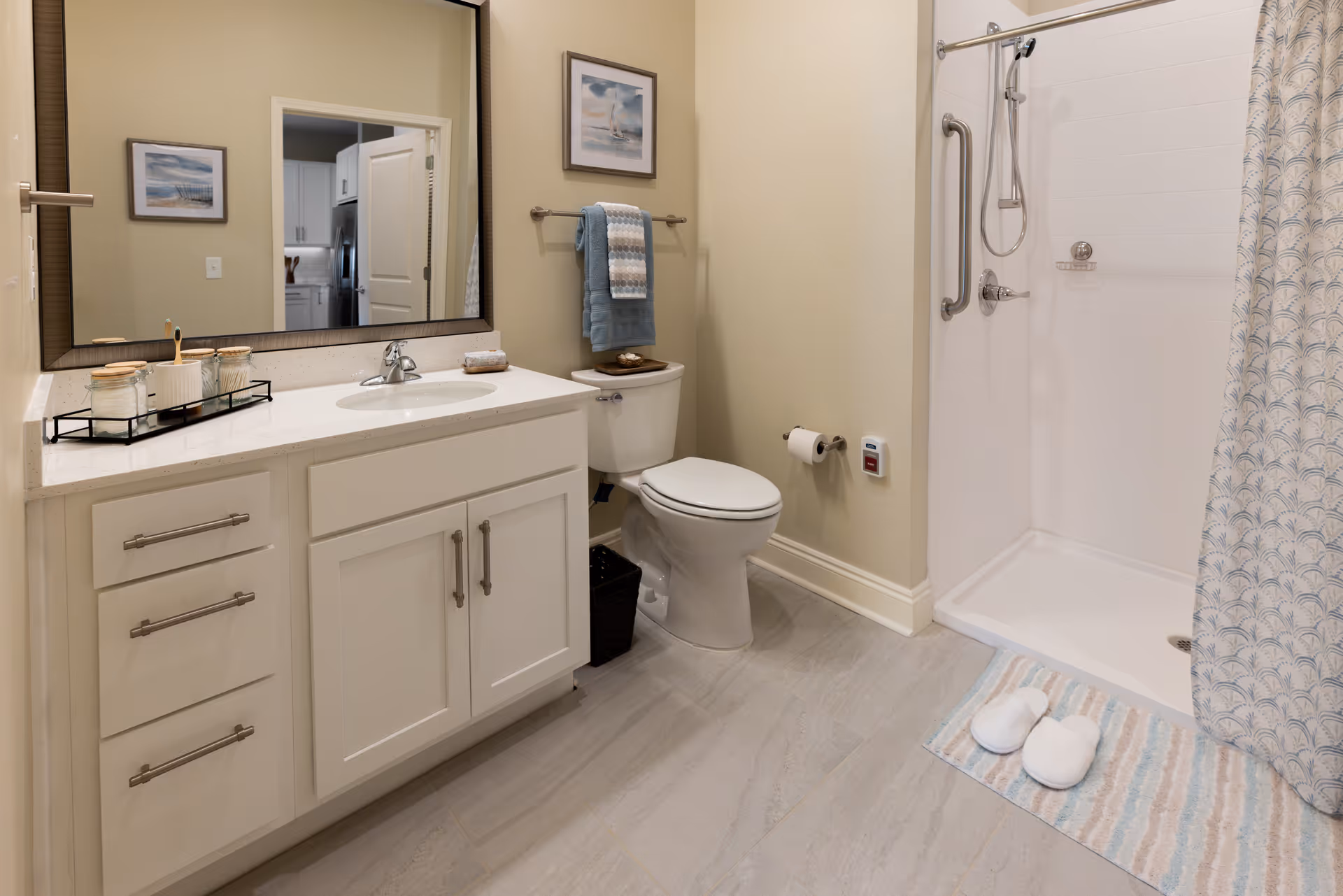 A clean and modern bathroom featuring a white vanity with a sink, a large mirror above it, a toilet with a towel rack and folded towels above, and a walk-in shower with a grab bar and a patterned shower curtain. A striped bath mat with white slippers is placed in front of the shower.