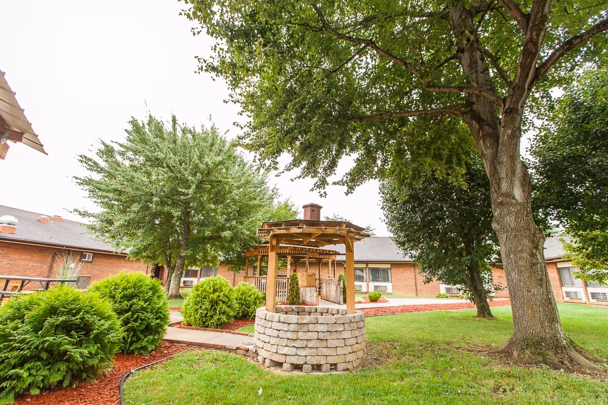 Outdoor garden area at Signature HealthCARE of Greeneville featuring a stone well with a wooden roof, surrounded by green grass, trees, and shrubs, with a brick building in the background.