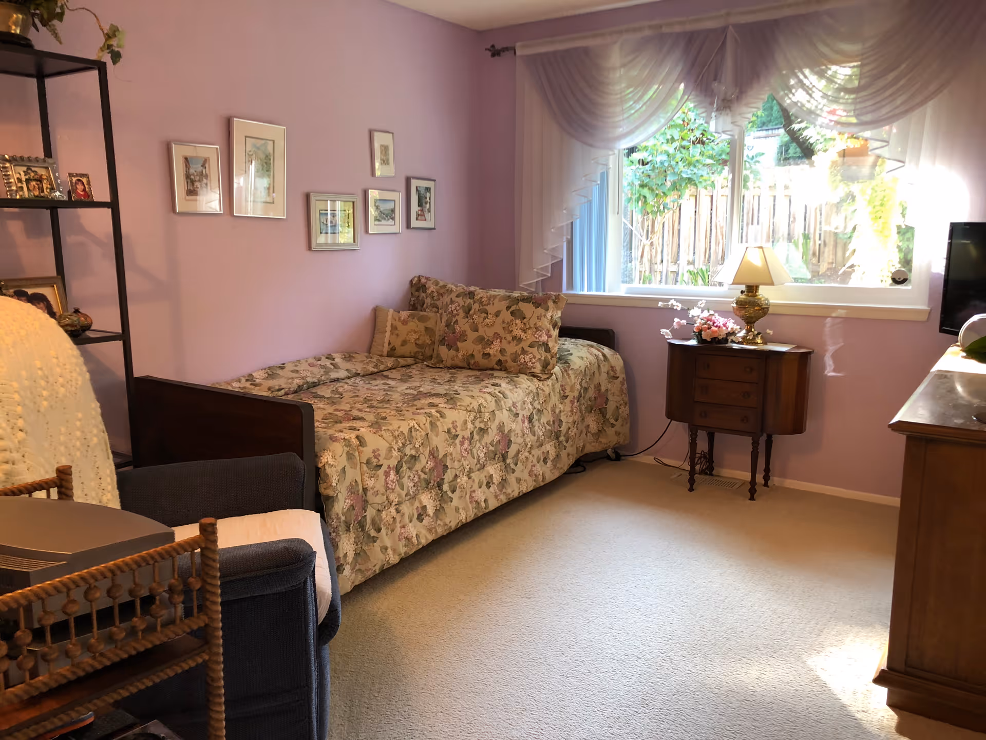 A cozy bedroom with lavender walls featuring a single bed with floral bedding and matching pillows. Next to the bed is a small wooden nightstand with a lamp and a flower arrangement. A large window with sheer white curtains lets in natural light and shows greenery outside. There is a dark armchair with a white knitted throw and a wooden shelving unit with framed photos and decorative items on the left side of the room. A wooden dresser is partially visible on the right side.
