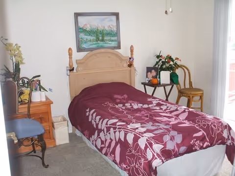 A small bedroom with a single bed covered in a purple and white floral bedspread. The bed has a wooden headboard. To the left of the bed is a wooden nightstand with a plant and other small items on it. There is a blue cushioned chair with a metal frame next to the nightstand. On the right side of the bed, there is a small table with a framed photo, a plant, and a small pumpkin decoration. A wooden chair is placed next to the table. A painting of a mountain landscape hangs on the wall above the bed. A window or glass door with vertical blinds is visible on the right side of the room.
