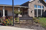 Exterior view of a building with stone and siding walls, large windows, and a covered entrance. In front of the building is a garden area with colorful flowers and a sign that reads 'Martin Health Center.'
