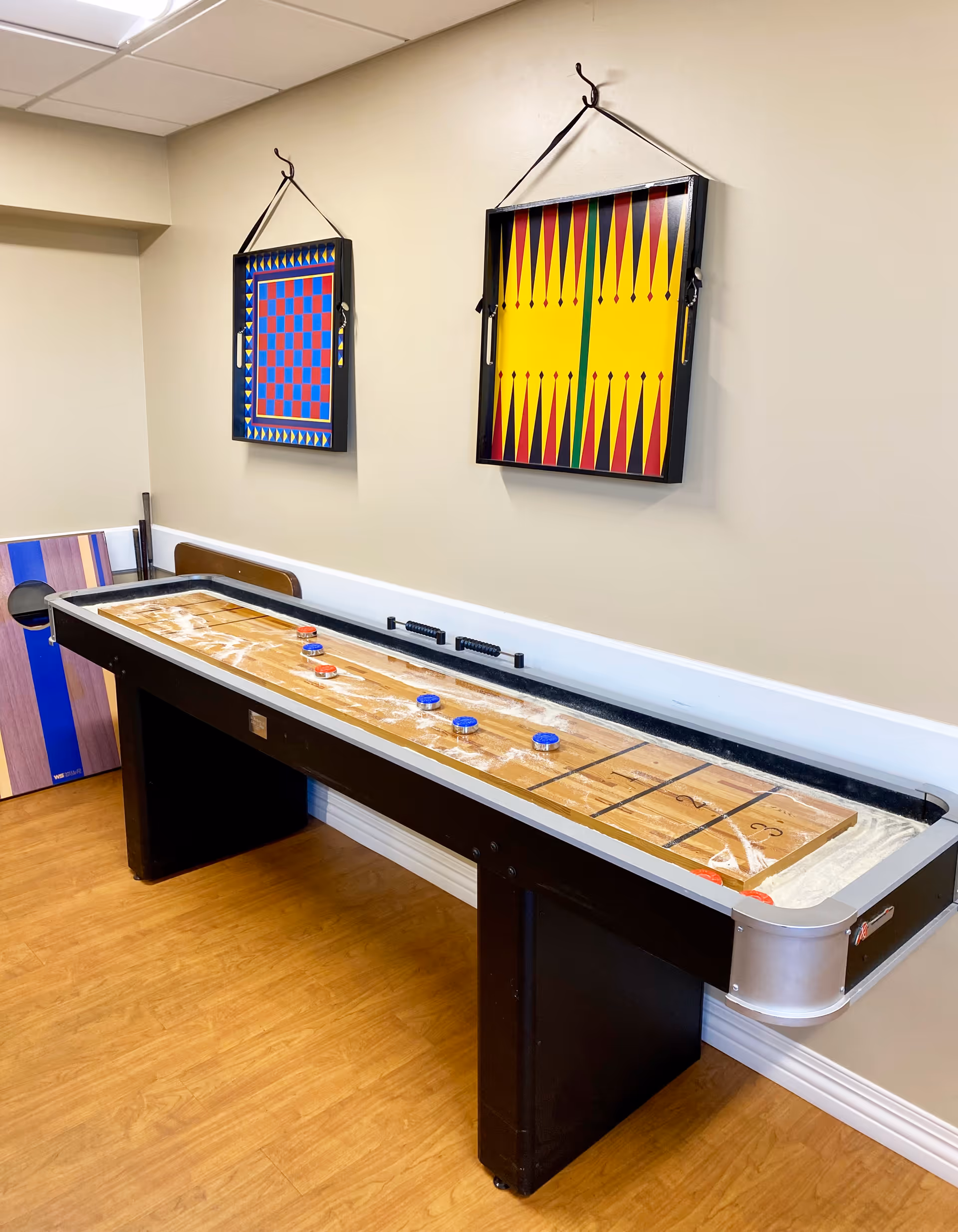 A shuffleboard table with pucks on it in a room with light-colored walls and wooden flooring. Two colorful game boards, one for checkers and one for backgammon, are hanging on the wall above the shuffleboard table. A wooden cornhole board is leaning against the wall in the corner.