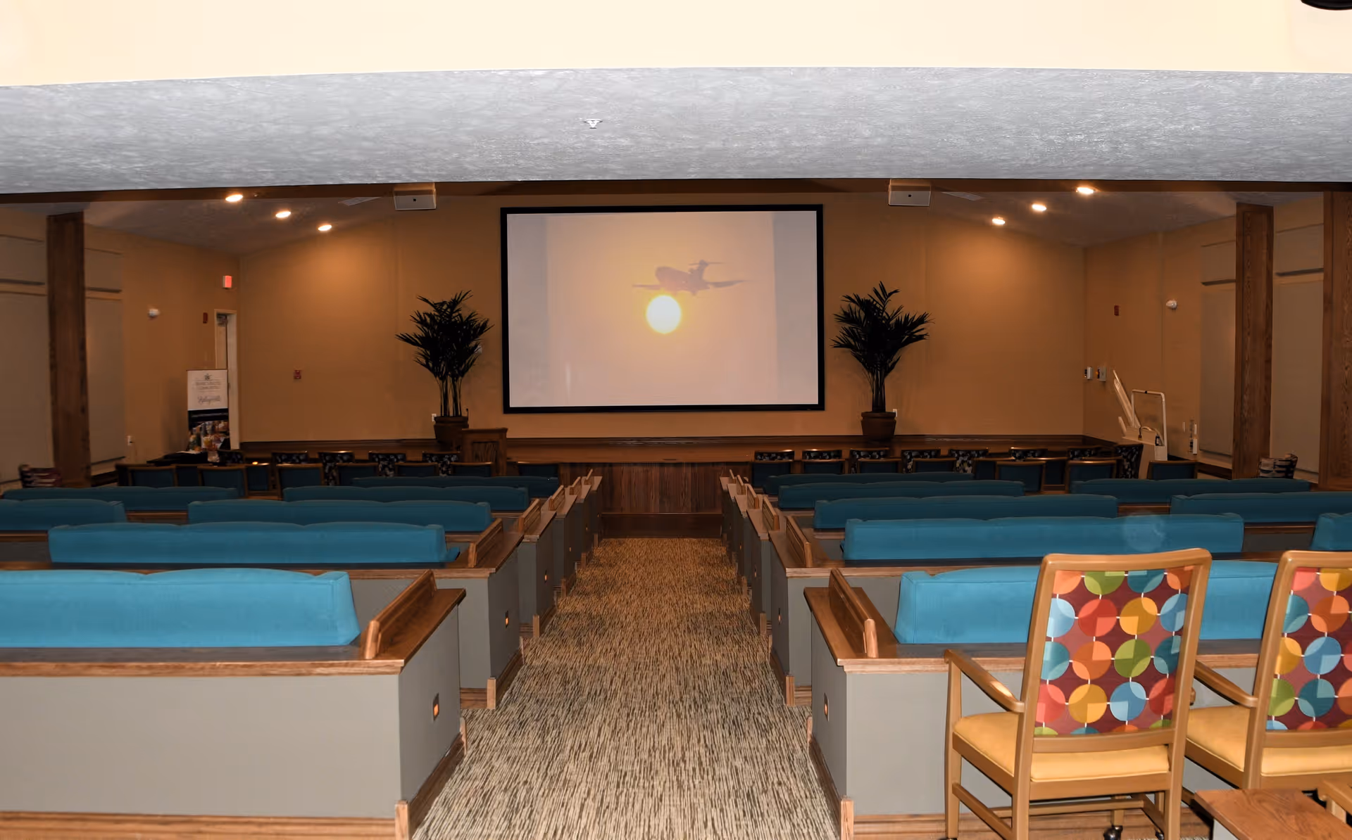 An auditorium-style room with rows of teal cushioned benches and colorful chairs facing a large projection screen displaying an image of an airplane flying in front of the sun. The room has beige walls, carpeted floor, and two potted plants on either side of the screen.