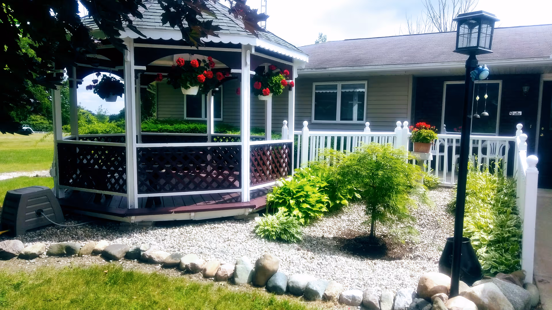 Landscaped front yard featuring a white gazebo with hanging flowers, gravel beds and plants in front of a single-story building.