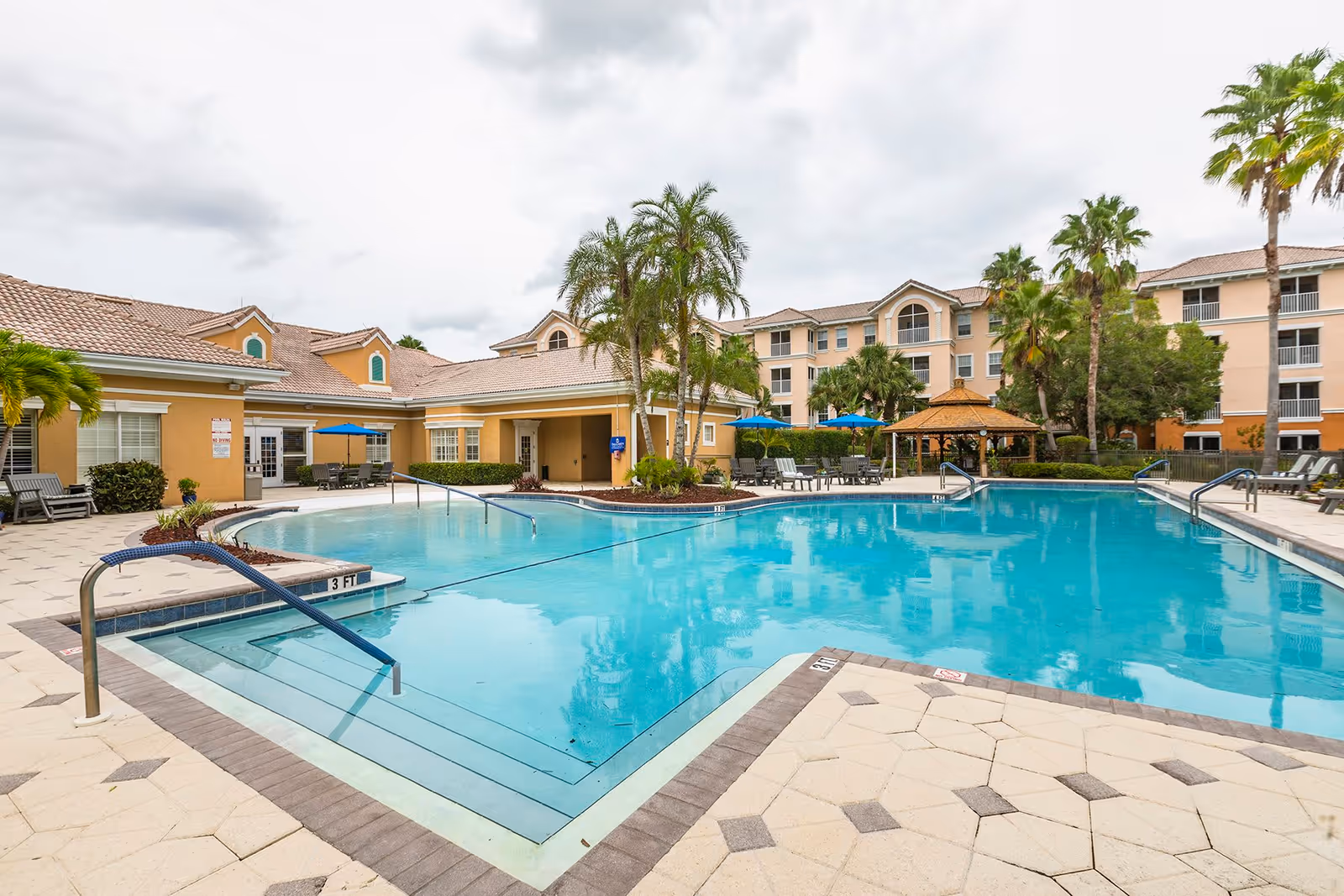 Outdoor swimming pool area at Aston Gardens At Pelican Pointe with clear blue water, surrounded by a tiled deck with lounge chairs, umbrellas, palm trees, and a gazebo. The building of the facility is visible in the background under a cloudy sky.