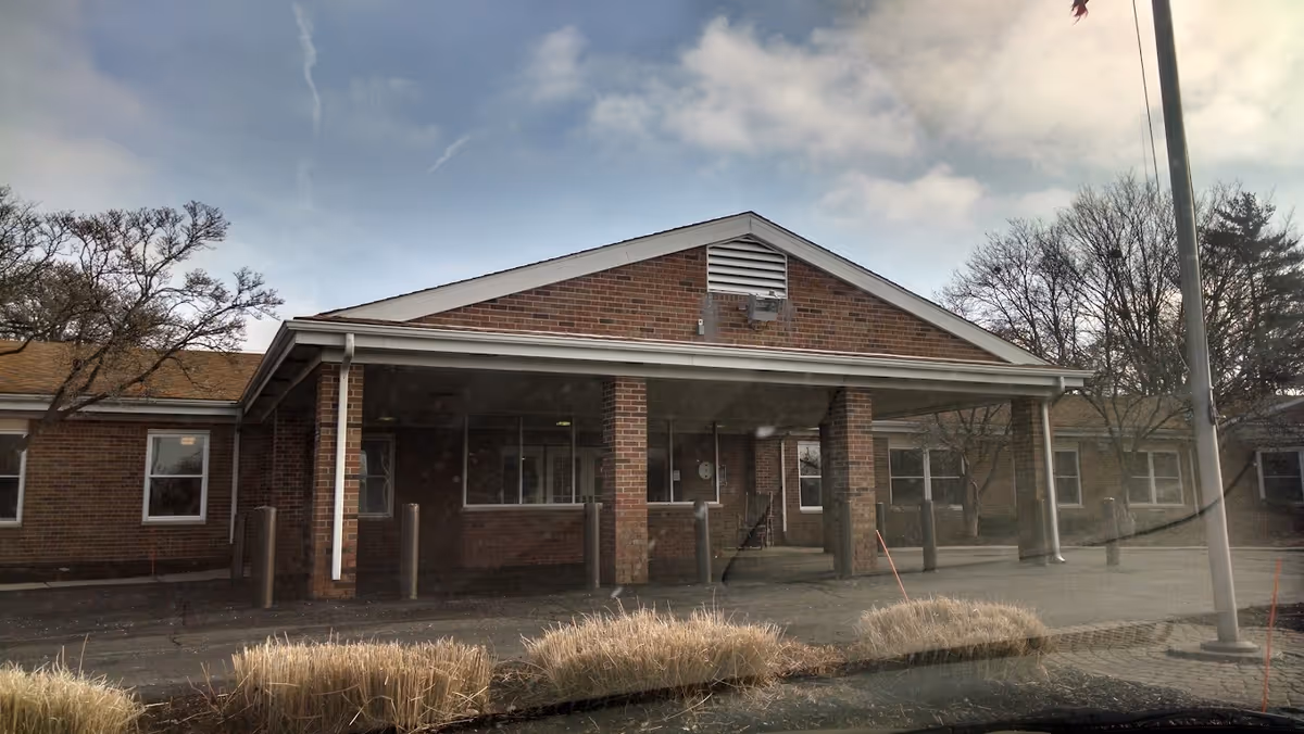 Exterior view of a single-story brick building with a covered entrance supported by brick columns. There are several windows along the building and some leafless trees around it. The sky is partly cloudy.