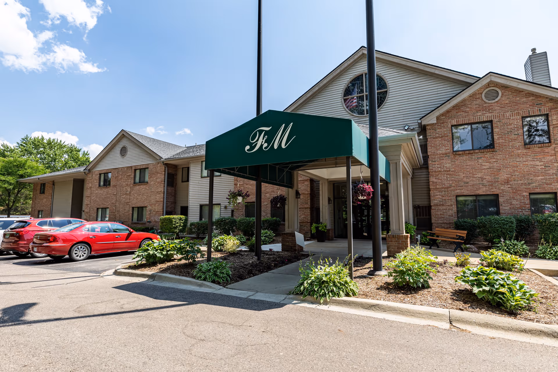 Exterior view of a senior living facility building with a green canopy entrance labeled 'FM'. The building has brick and siding walls, multiple windows, and a circular window with an American flag visible inside. There are parked cars and landscaped plants around the entrance under a clear blue sky.