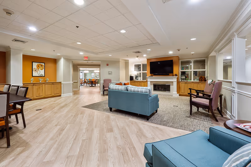 A spacious and well-lit living room area in a senior living facility featuring light wood flooring, a blue sofa set around a carpeted area, a fireplace with a mounted flat-screen TV above it, wooden cabinets on either side of the fireplace, and several chairs. The walls are painted in neutral tones with some orange accent walls and decorative artwork. In the background, a dining area with tables and chairs is visible through an open doorway.