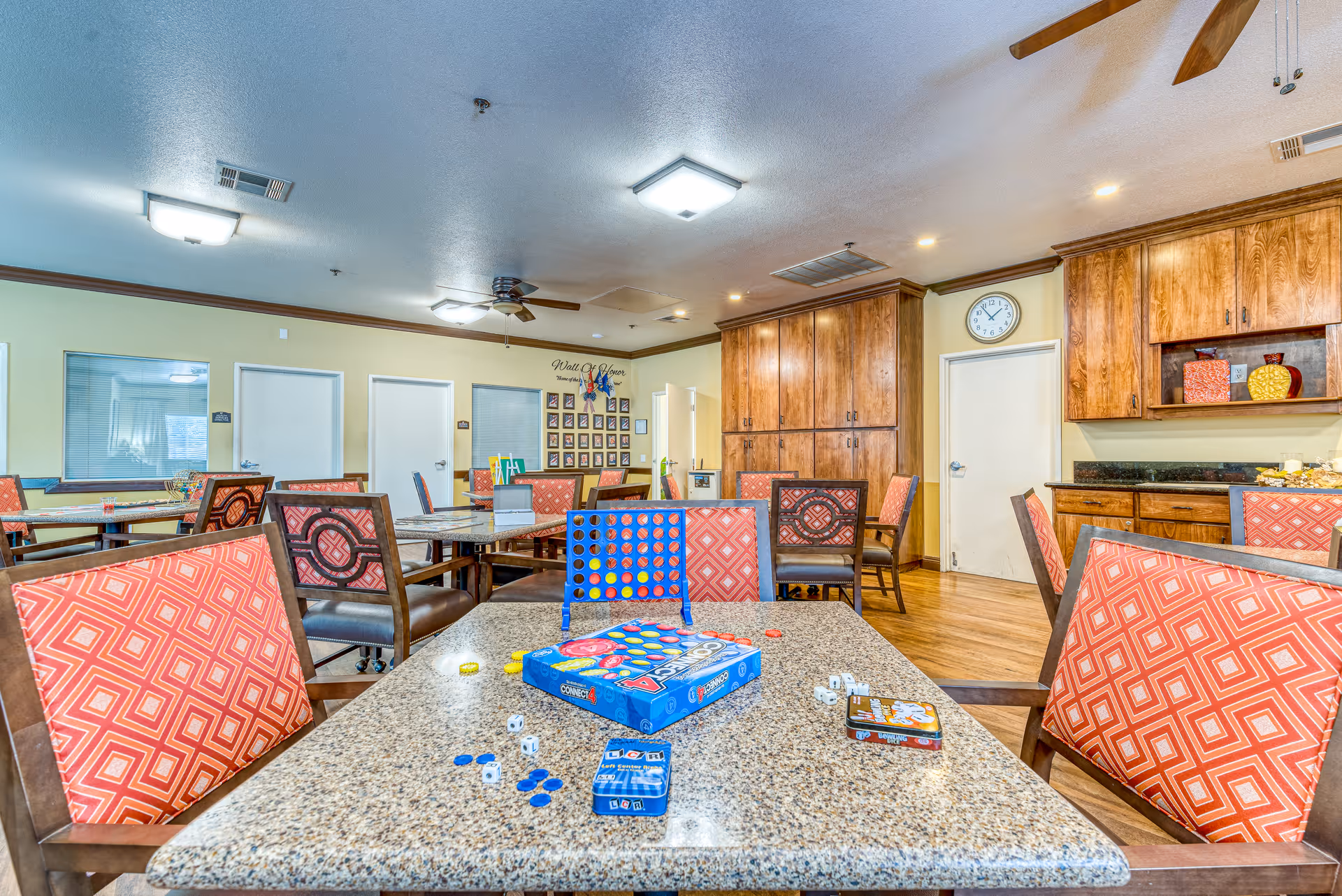 A bright and spacious common activity room with multiple tables and chairs featuring red patterned upholstery. On the nearest table, there are board games including Connect 4, dice, and game cards. The room has wooden cabinets, a clock on the wall, ceiling fans, and overhead lighting. The walls are light yellow with some framed photos and a 'Wall of Honor' display.