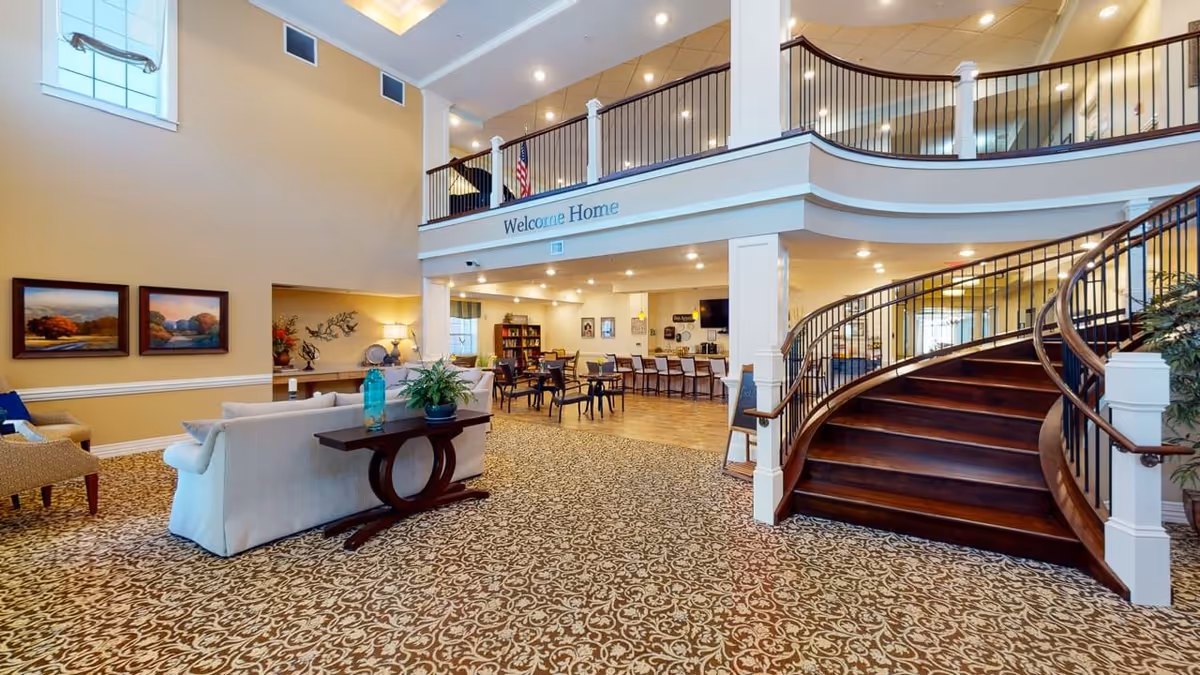 Spacious senior living facility lobby with a curved wooden staircase, seating area, and a 'Welcome Home' sign on the upper balcony.