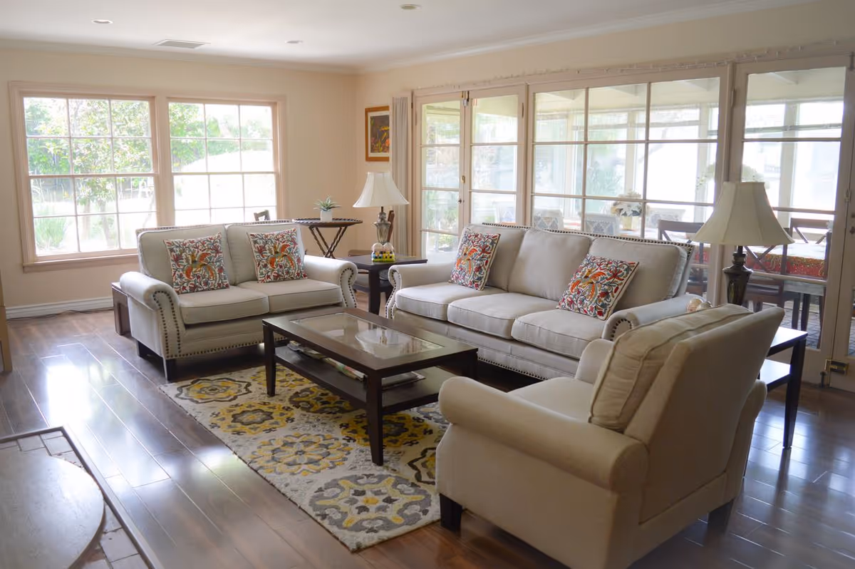 A bright and cozy living room with three beige upholstered sofas adorned with colorful patterned throw pillows. A glass-top coffee table sits on a decorative rug with yellow, gray, and white patterns. The room has large windows and glass doors that let in natural light and offer a view of an outdoor patio area with a dining table and chairs. Two table lamps with white shades are placed on side tables beside the sofas.