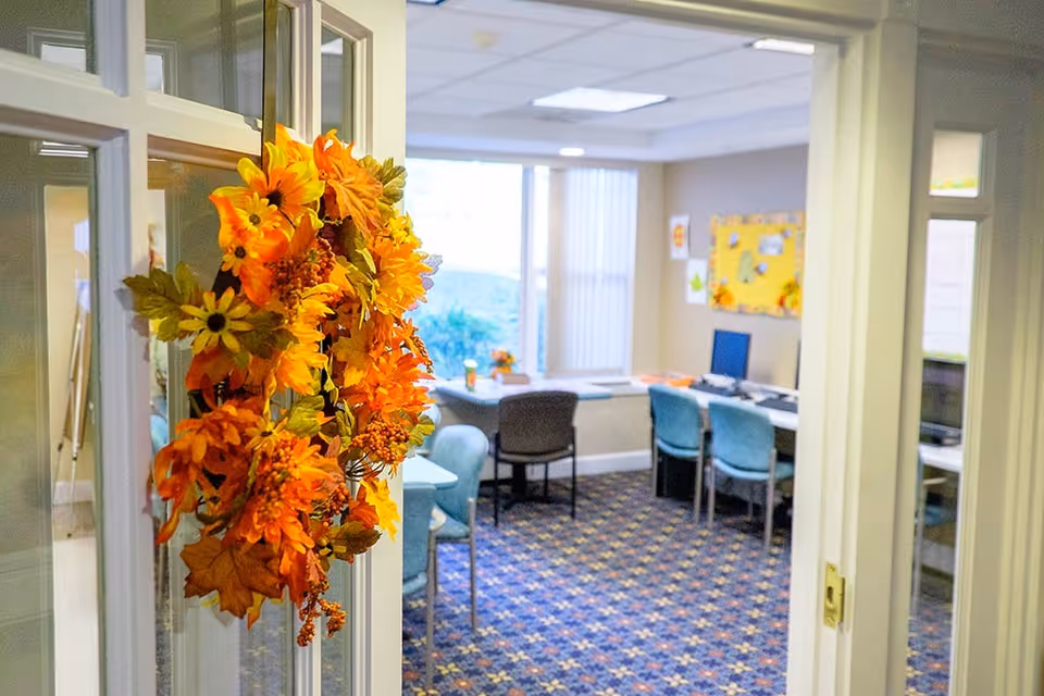 View through a glass door decorated with an autumn-themed wreath into a room with desks, chairs, computers, and a bulletin board, suggesting a communal or activity area.