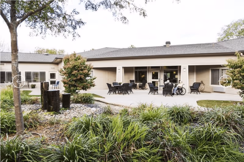 Outdoor patio area of a single-story assisted living facility with several black chairs and tables arranged on a concrete surface. Two people are seated, one in a wheelchair, enjoying the outdoor space. The patio is surrounded by greenery, including bushes, grass, and a tree, with a small rock garden and wooden posts in the foreground.