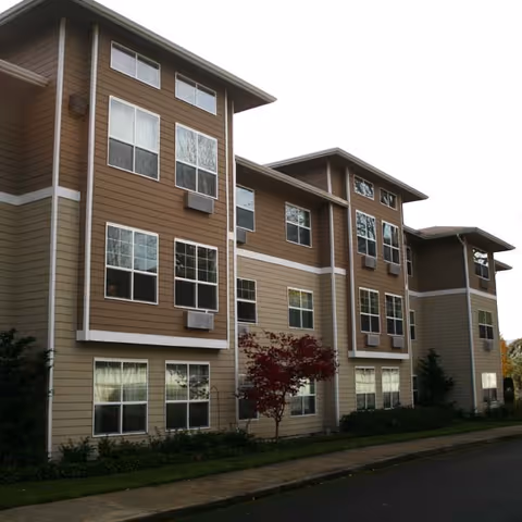 Three-story beige and brown apartment-style building with many windows and small landscaping along a sidewalk.