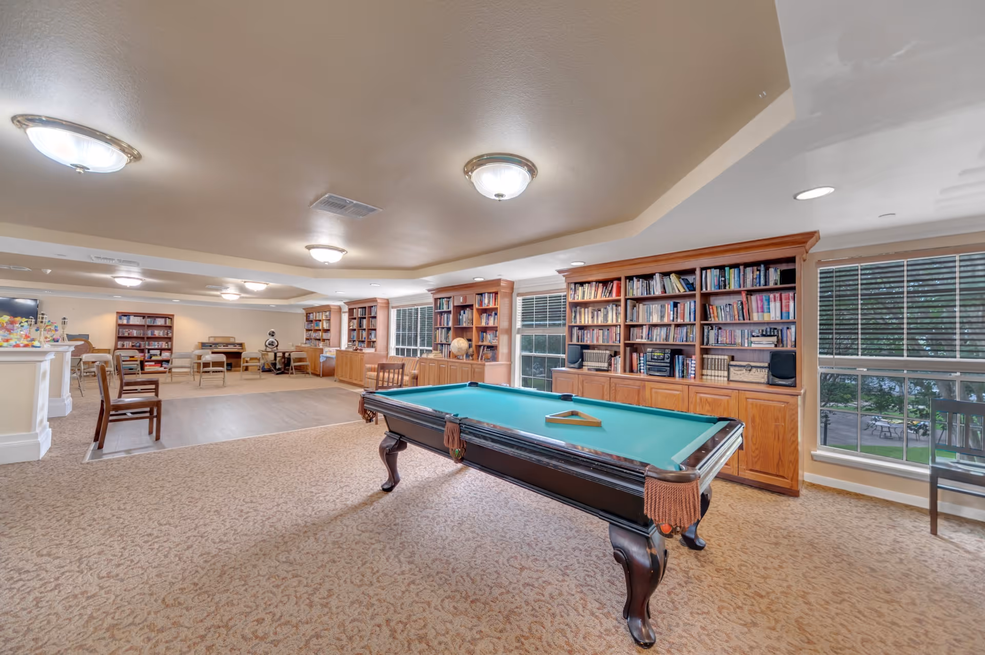 A spacious recreational room with a pool table in the center, surrounded by carpeted flooring. The room features large wooden bookshelves filled with books along the walls, several windows with blinds letting in natural light, and multiple ceiling lights. There are chairs and tables arranged in the background, creating a comfortable and inviting atmosphere.