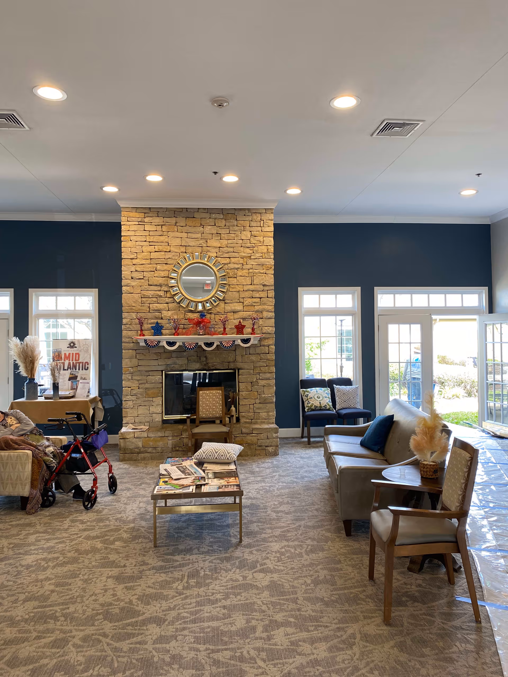 A cozy senior living facility common area with a stone fireplace in the center, decorated with patriotic items and a round mirror above it. The room has blue walls, large windows, and glass doors letting in natural light. There are several chairs and sofas arranged around a coffee table with newspapers on it. A walker is visible next to one of the chairs.