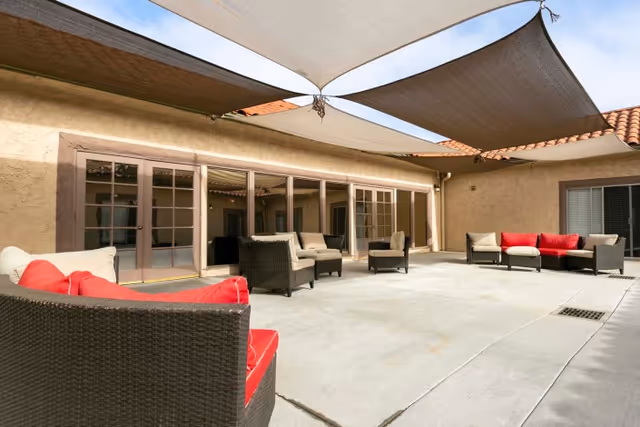 Outdoor courtyard with wicker lounge chairs and sofas with red cushions beneath triangular shade sails in front of glass doors.