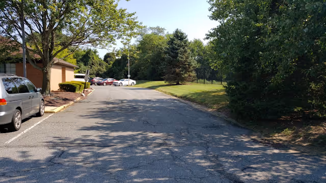 A paved driveway or parking area next to a brick building with several parked cars on the left side. The area is surrounded by green trees and grass under a clear blue sky.