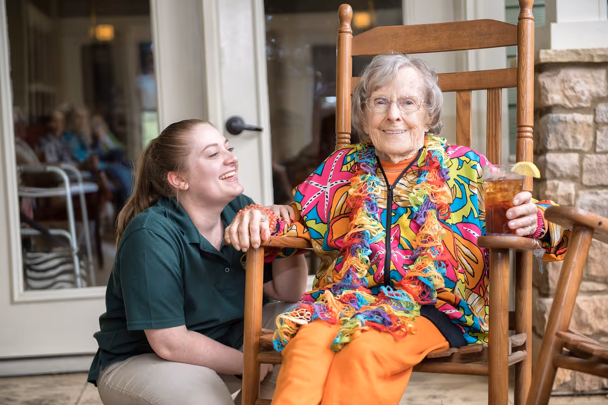 An elderly woman wearing a colorful outfit and a rainbow scarf sits in a wooden rocking chair holding a glass of iced tea with a lemon wedge. A young woman in a green shirt kneels beside her, smiling and holding her hand. They are on a patio area with a glass door and stone wall in the background.