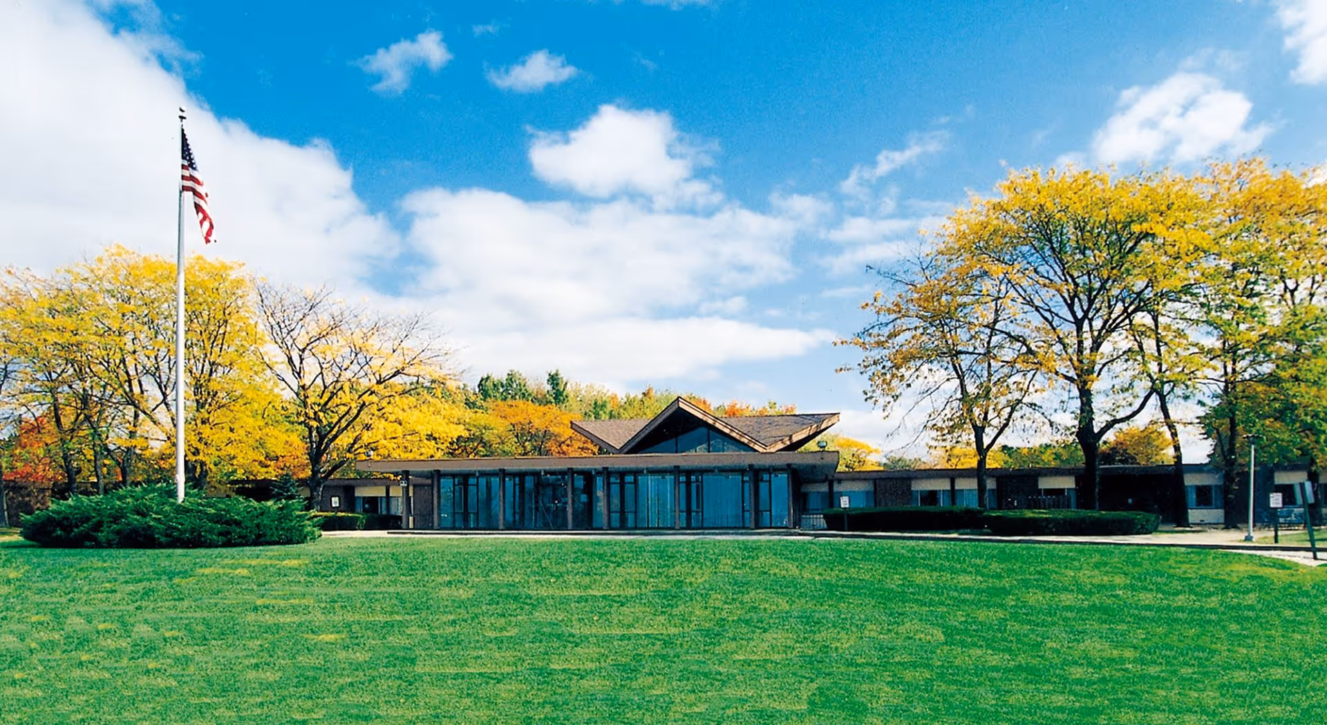 Front exterior of a single-story nursing facility with a wide green lawn, an American flag, and trees under a blue sky.