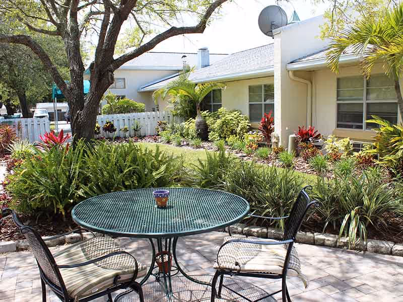Outdoor patio with a round metal table and three chairs surrounded by landscaped garden and a single-story building.