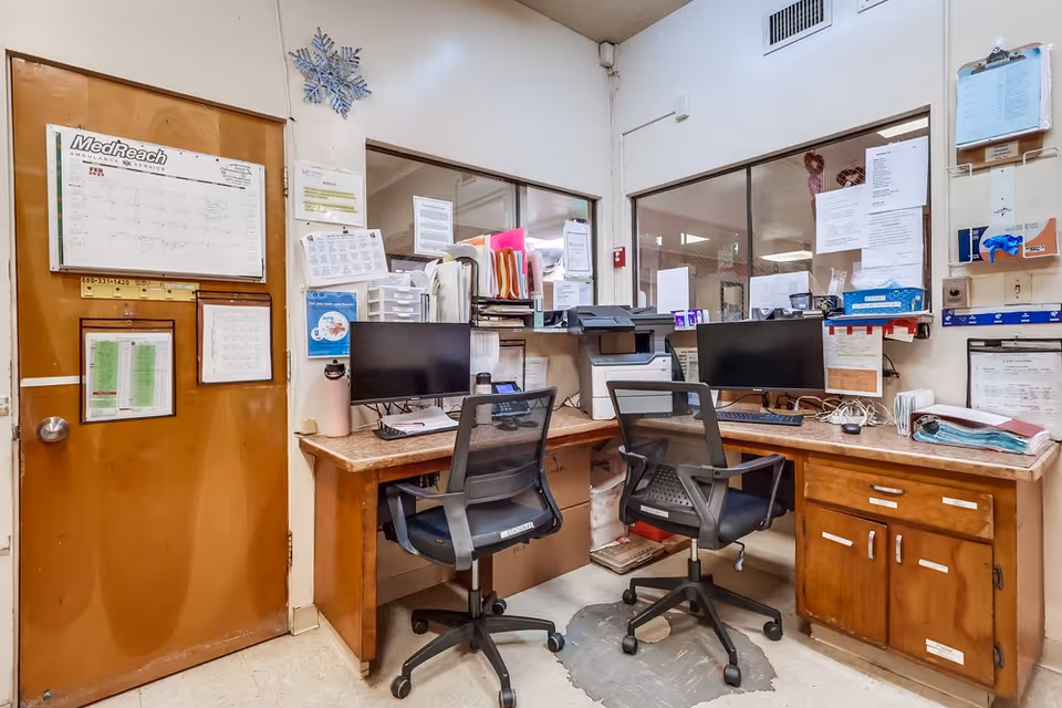 An office area with two black mesh office chairs in front of a wooden desk with two computer monitors, a printer, and various papers and files. The room has beige walls, a wooden door with posted schedules and notices, and windows looking into another room.
