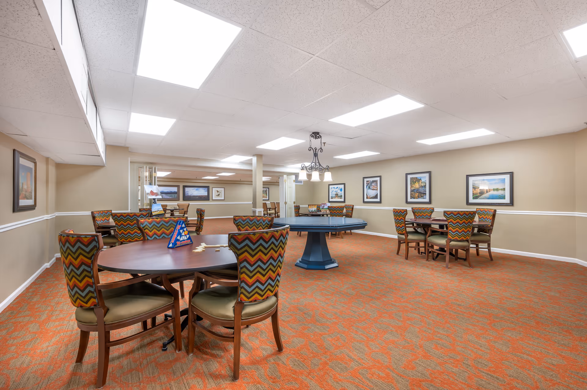 Spacious, brightly lit communal dining/activity room with round tables and chevron-patterned chairs and framed artwork on the walls.