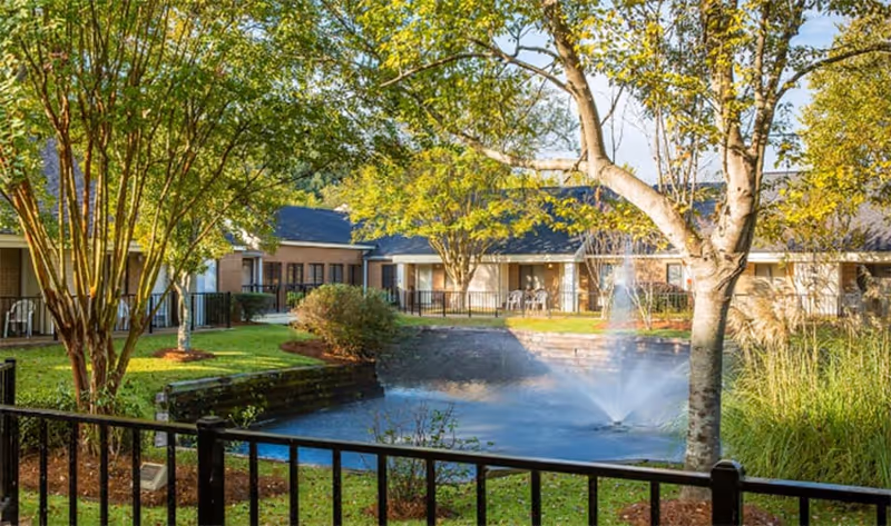 Outdoor view of The Stiles Apartments featuring a small pond with a water fountain, surrounded by trees, bushes, and a black metal fence. Apartment buildings with patios and chairs are visible in the background under a clear sky.