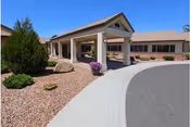 Exterior view of a single-story building with a covered entrance supported by columns, surrounded by desert landscaping with rocks, shrubs, and a small tree under a clear blue sky.