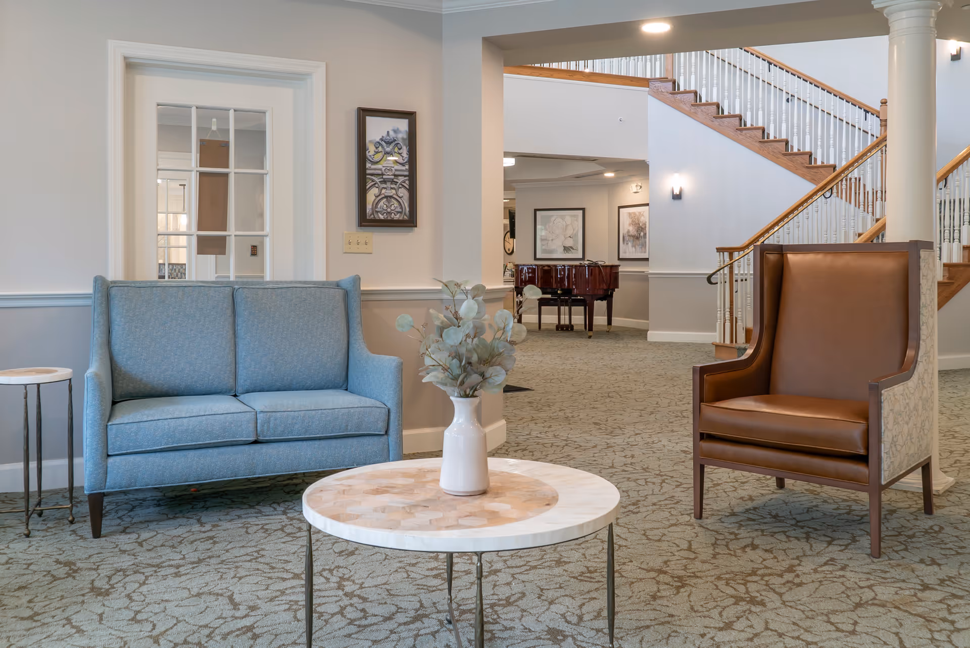 A cozy sitting area in a senior living facility with a blue loveseat, a brown leather armchair, and a round table with a vase of greenery. In the background, there is a grand piano, framed artwork on the walls, and a staircase with wooden handrails and white balusters.