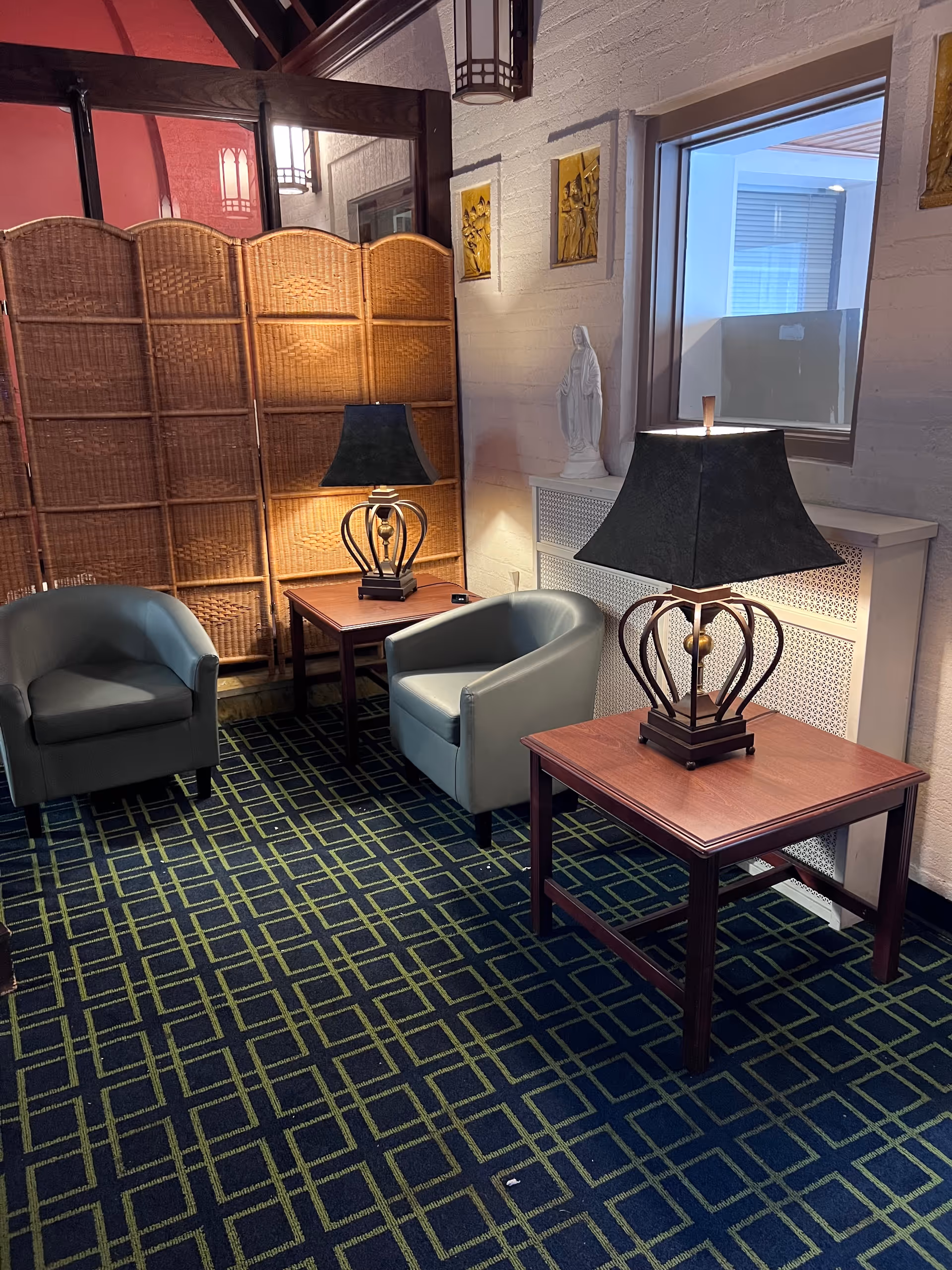 A cozy seating area with two gray armchairs and two wooden side tables, each with a decorative lamp. The floor is covered with a patterned carpet in dark blue and green. Behind the seating area is a woven room divider and a white wall with religious artwork and a statue. A window is visible on the right side.
