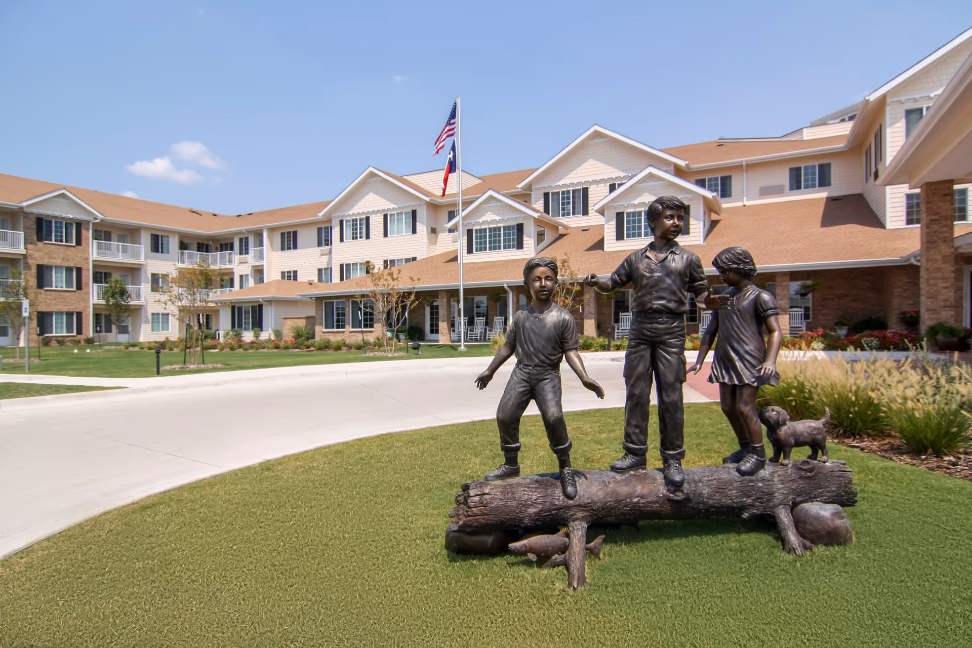 Outdoor view of Highland Estates - Sky Active Living facility with a bronze statue of three children and a dog playing on a log in the foreground. The building has multiple stories with beige siding, white trim, and a tan roof. An American flag is flying on a flagpole near the entrance.