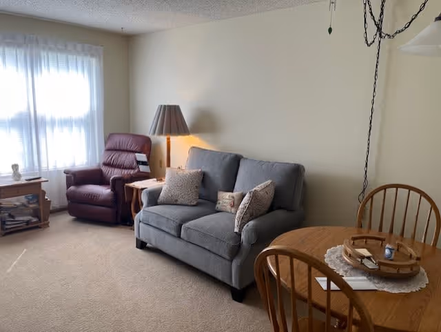A cozy living room area with a gray upholstered sofa adorned with three patterned pillows, a burgundy recliner chair next to a wooden side table with a lamp, a wooden round dining table with chairs, and a window with sheer white curtains allowing natural light to fill the room.