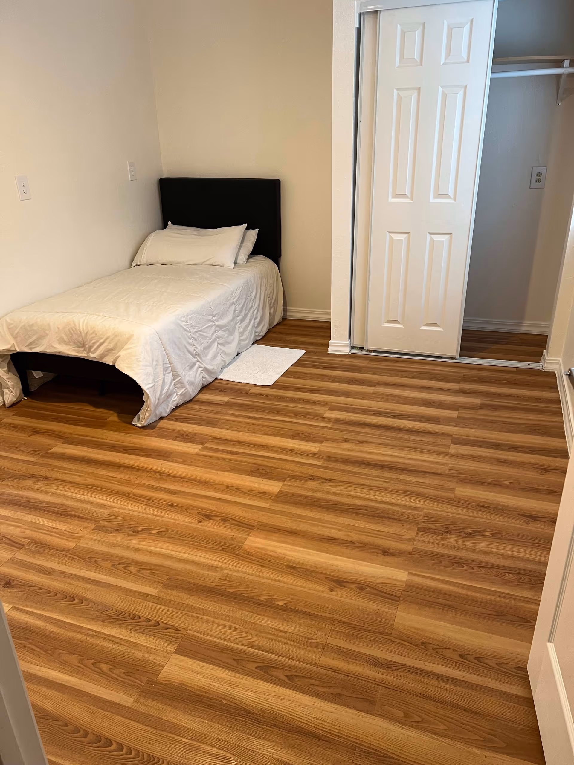 A small bedroom with wood laminate flooring, a single bed with white bedding and pillows against the left wall, and a closet with sliding white doors on the right side.