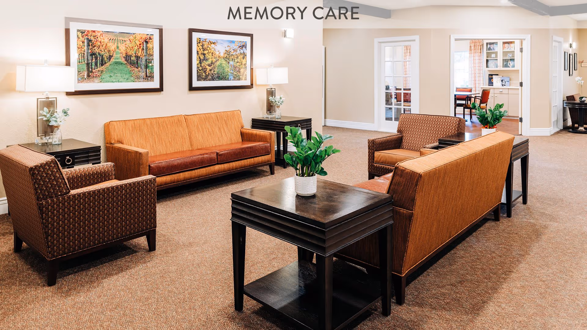 A cozy memory care lounge area with two orange sofas and two patterned armchairs arranged around a dark wooden coffee table with a potted plant. The room has beige walls, carpeted floor, two framed landscape paintings on the wall, and side tables with lamps and small plants. In the background, there are glass doors leading to another room with more seating.