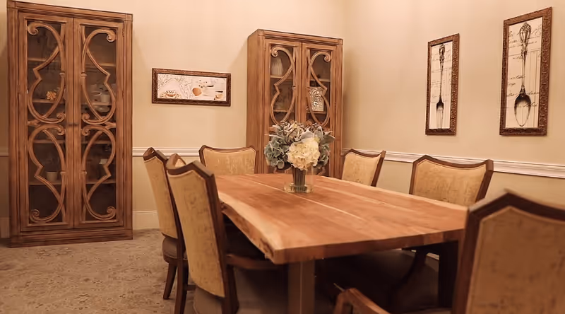 Wooden dining table with a floral centerpiece surrounded by upholstered chairs and flanked by two decorative wooden cabinets in a warmly lit dining room.