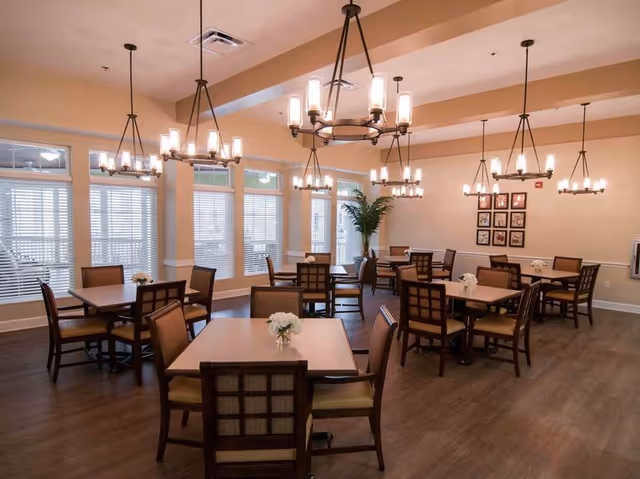 A well-lit dining room with several square wooden tables and matching chairs arranged neatly. Each table has a small floral centerpiece. Large windows with white blinds line the walls, and multiple modern chandeliers hang from the ceiling. The room has a warm, inviting atmosphere with light-colored walls and wooden flooring.