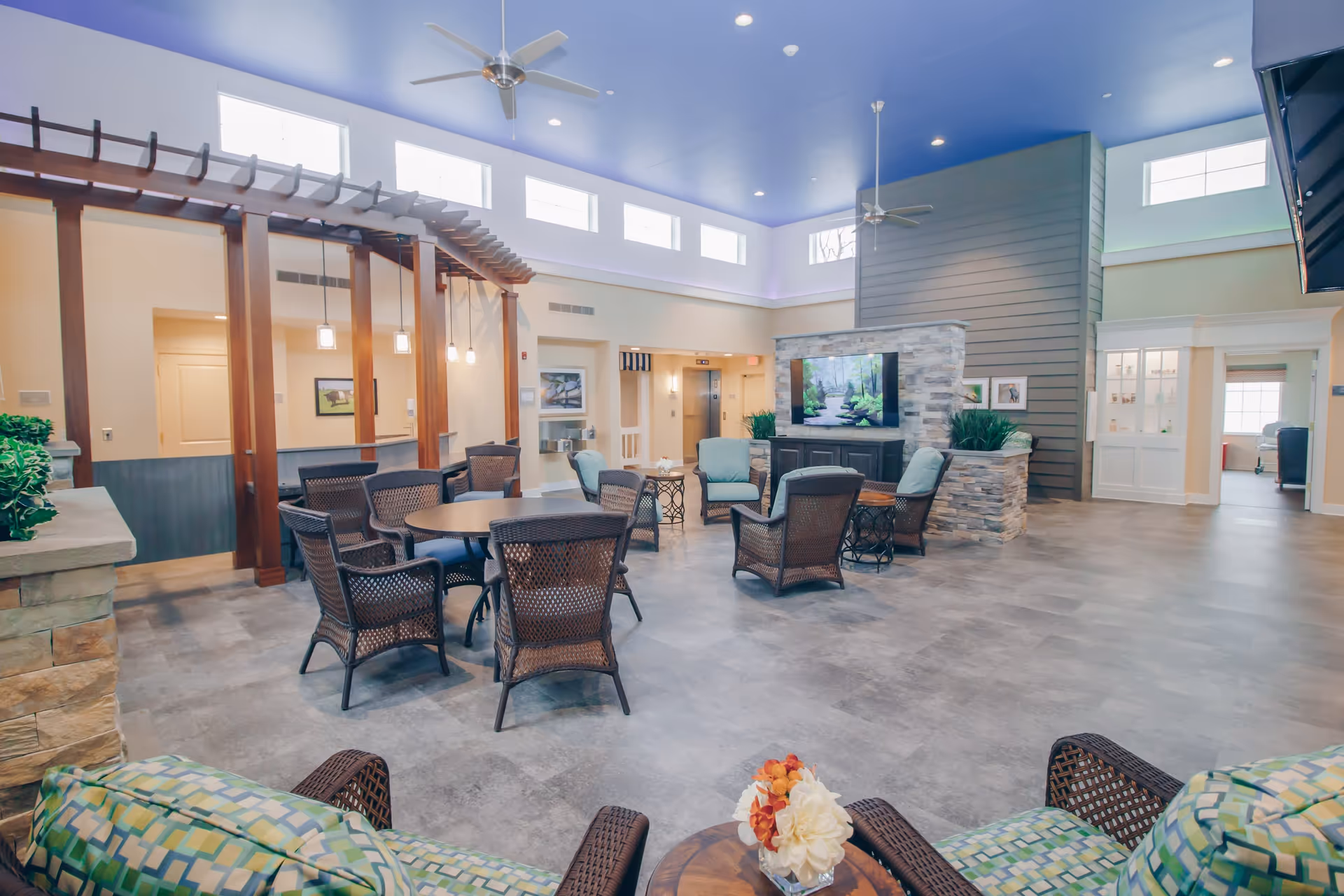 A spacious senior living common area with multiple wicker chairs and tables arranged around a stone fireplace with a mounted TV. The room has a high blue ceiling with ceiling fans, large windows near the ceiling allowing natural light, and a wooden pergola structure with hanging pendant lights. There are plants and framed pictures decorating the space.